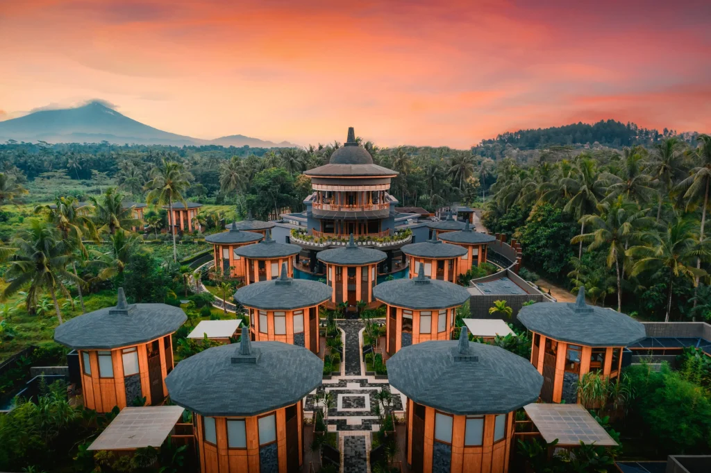 Aerial view of a circular villa resort surrounded by lush greenery and palm trees, with a volcano in the background during sunset.